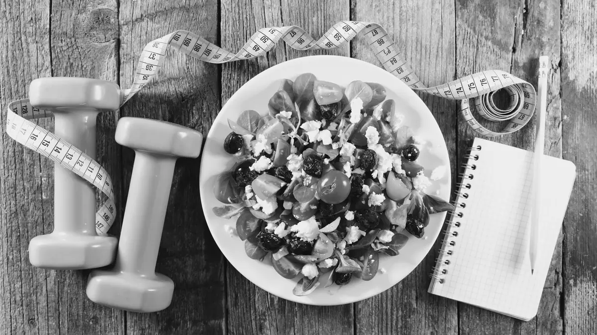 Un plato de ensalada, mancuernas y una cinta métrica sobre una mesa de madera